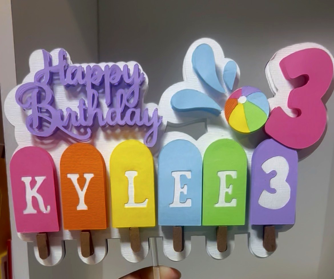 A handmade layered cake topper set spelling the name Kylee across six pastel popsicle shapes with a Happy Birthday script and a number three